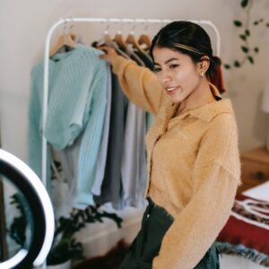 Trendy smiling woman taking clothes from rail while shooting video