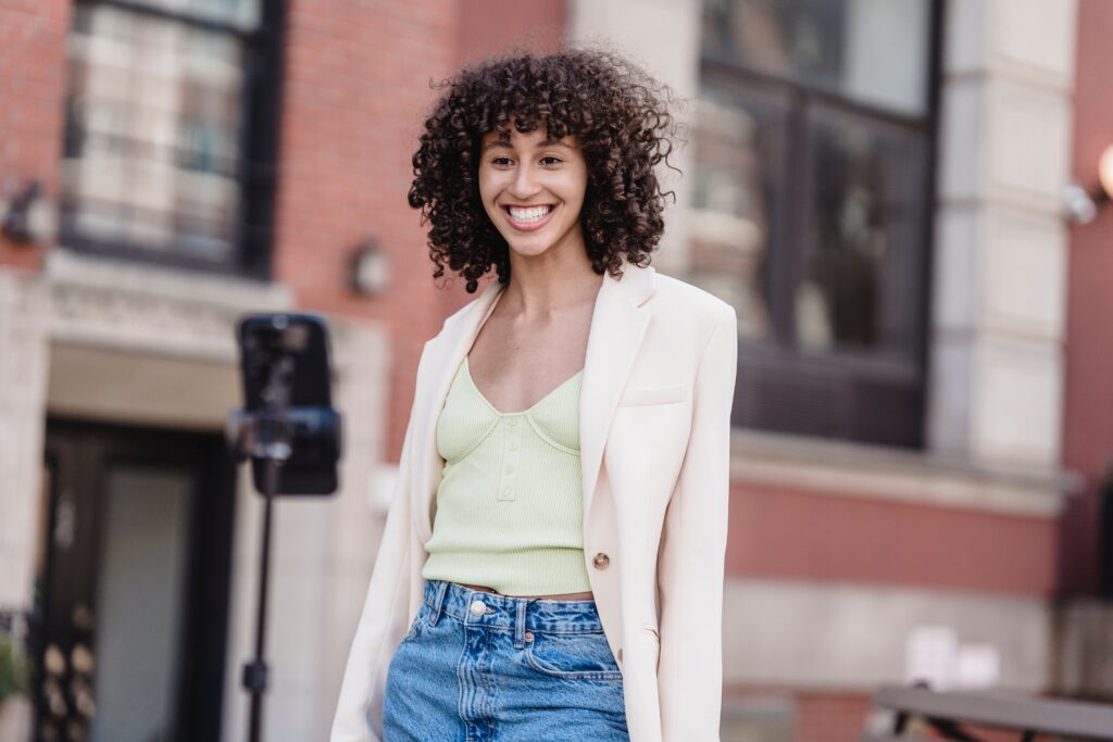 Glad ethnic female in white classic jacket and denim trousers chatting on video online on blurred background of urban building