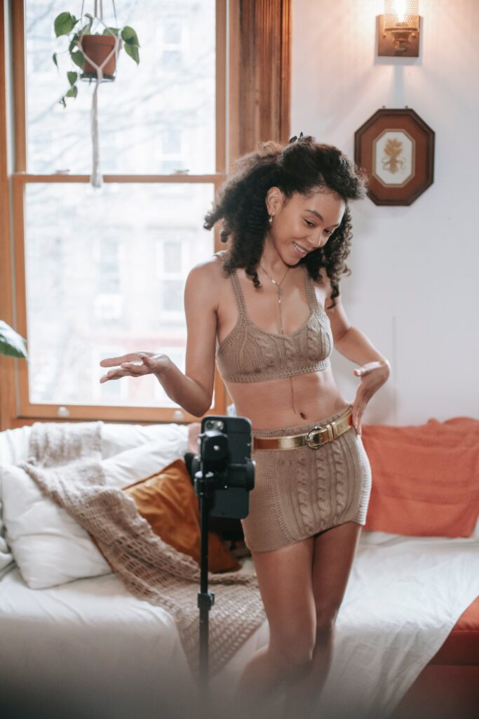 Positive ethnic female blogger promoting clothes for social media and filming video on cellphone on tripod
